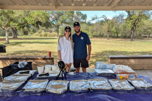 Two SH 130 staff members smiling behind a table of covered food trays at an outdoor community event.