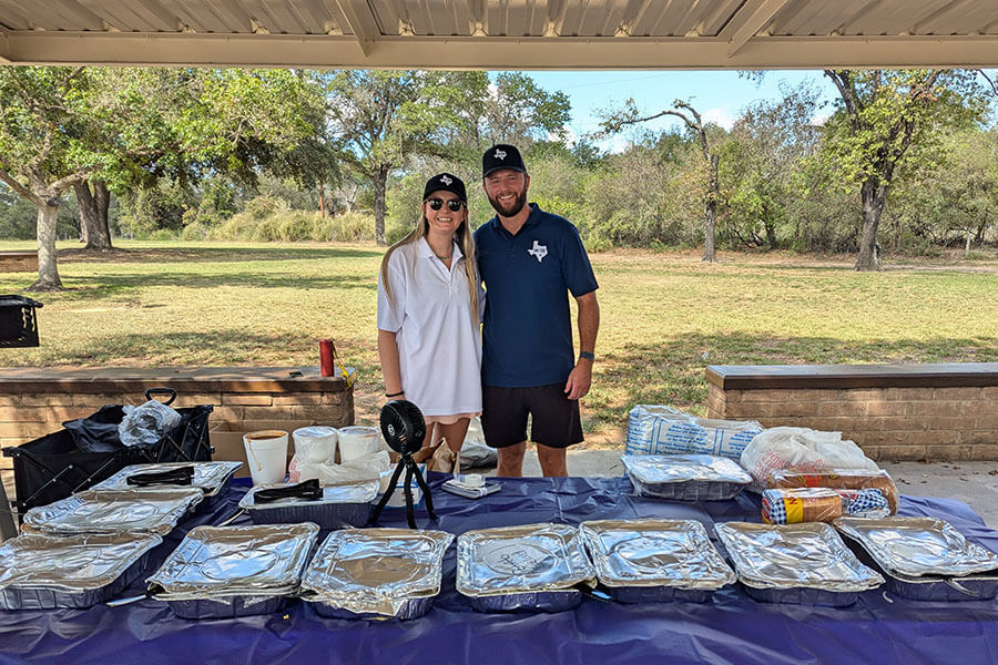 Two SH 130 staff members smiling behind a table of covered food trays at an outdoor community event.