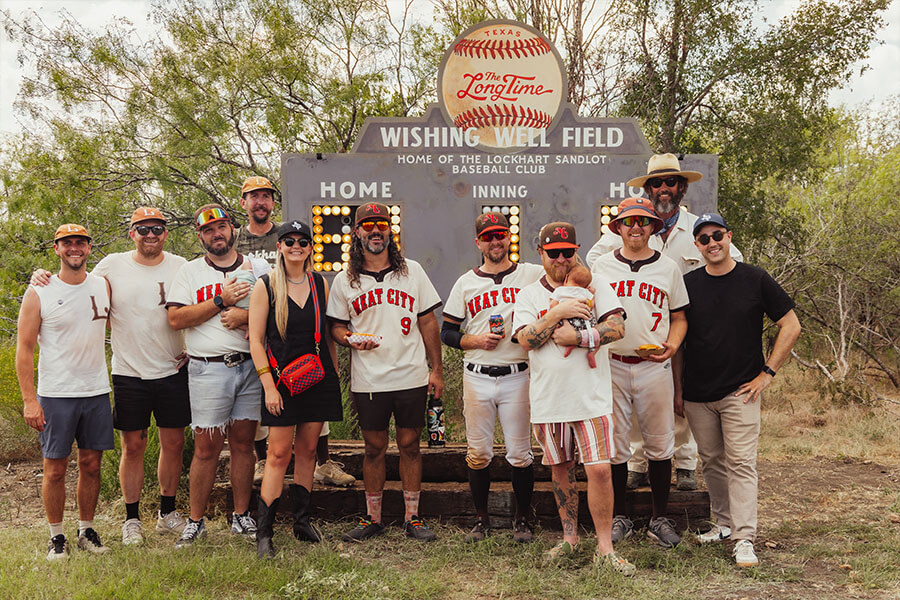 A group of people posing in front of the Wishing Well Field scoreboard, home of the Lockhart Sandlot Baseball Club, at a community event.