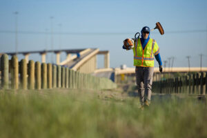 An SH 130 maintenance worker in a high-visibility vest carrying a weed trimmer along the highway roadside near an overpass.