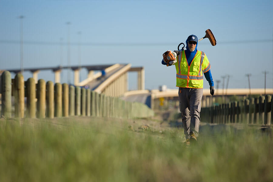 An SH 130 maintenance worker in a high-visibility vest carrying a weed trimmer along the highway roadside near an overpass.