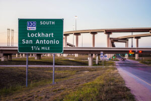A highway directional sign reading "130 South Toll – Lockhart, San Antonio – 1¼ Mile" with a multi-level interchange in the background at dusk.