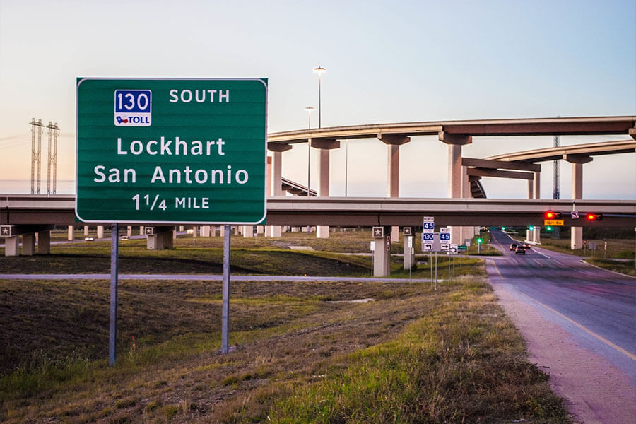 A highway directional sign reading "130 South Toll – Lockhart, San Antonio – 1¼ Mile" with a multi-level interchange in the background at dusk.