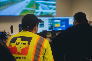 Two SH 130 employees monitoring highway traffic conditions from a control room with multiple screens displaying aerial views of the roadway.