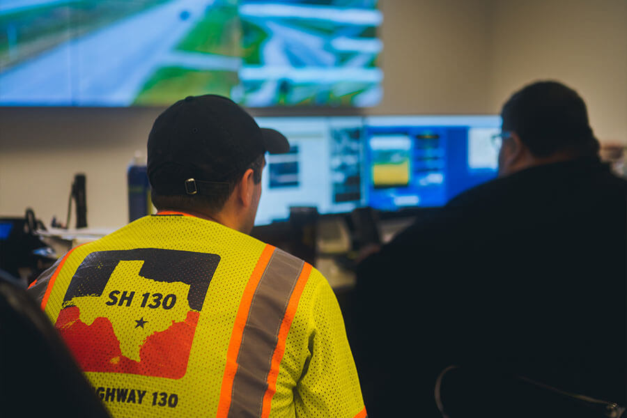 Two SH 130 employees monitoring highway traffic conditions from a control room with multiple screens displaying aerial views of the roadway.