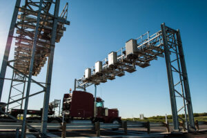 A commercial truck passing under an SH 130 electronic toll gantry on a clear day.