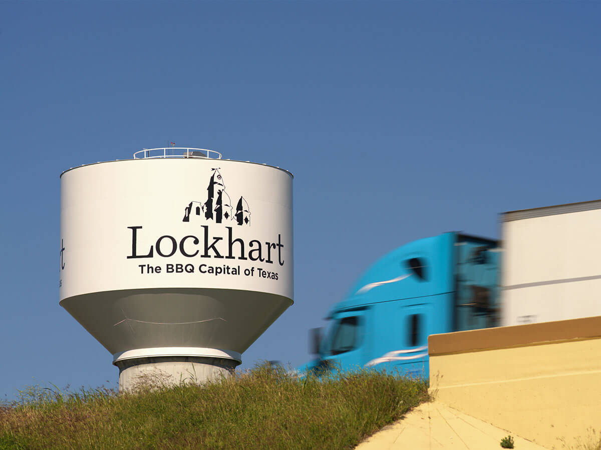 A commercial truck passes by the Lockhart water tower, reading "Lockhart – The BBQ Capital of Texas," under a clear blue sky.