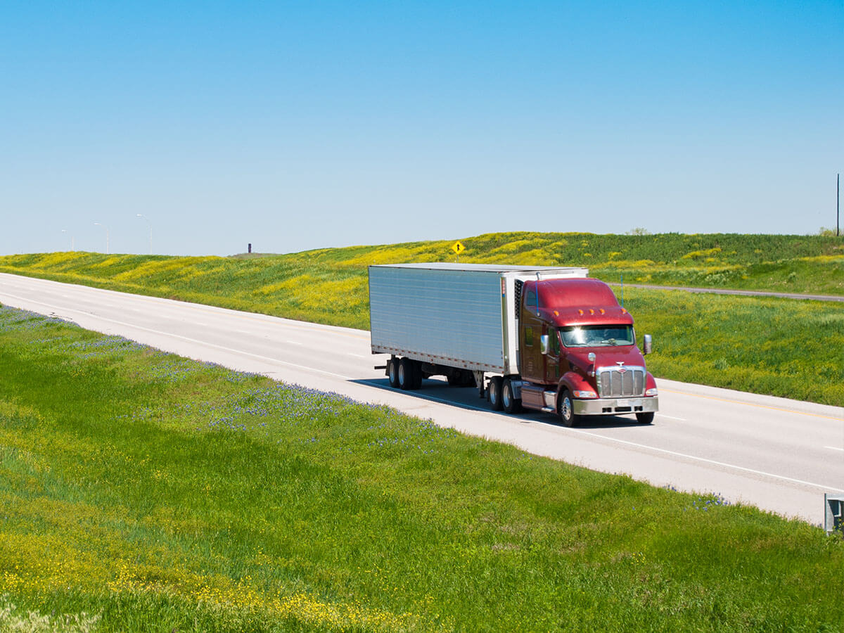 A semi-truck traveling along SH 130 past green fields and Texas wildflowers under a clear blue sky.