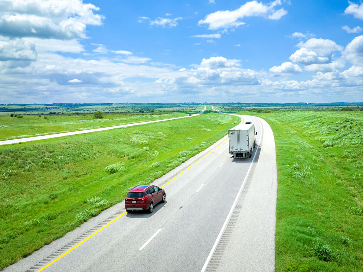 A red car and white box truck travel down an open stretch of SH 130 South, flanked by wide green fields under a clear cloudy sky.