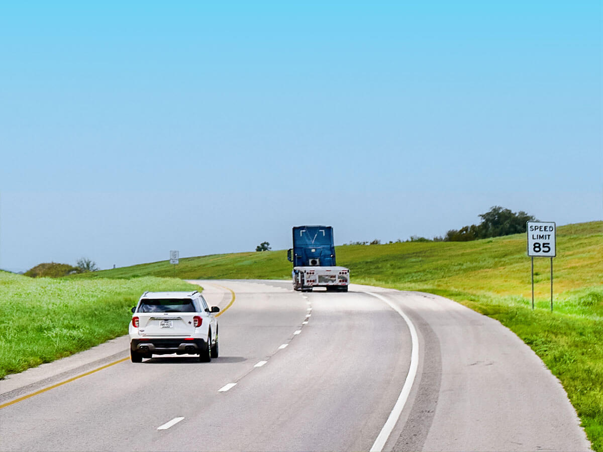A car and commercial truck traveling on a wide, open SH 130 highway with a speed limit sign alongside green rolling hills.