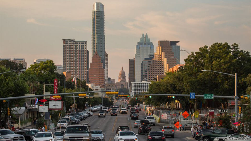 The downtown Austin skyline at dusk, with the Texas State Capitol dome visible behind traffic on South Congress Avenue.