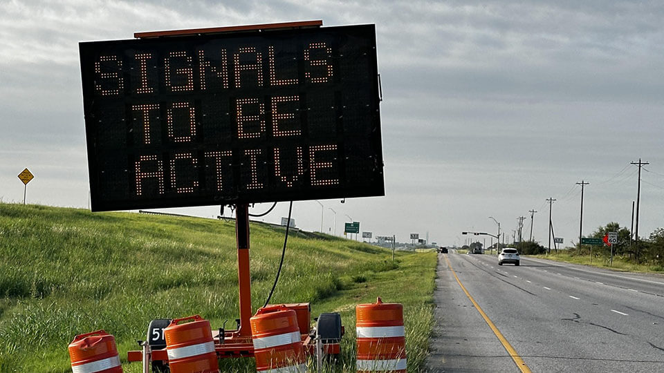 A portable digital roadside message board reading "Signals to Be Active," surrounded by orange traffic barrels.