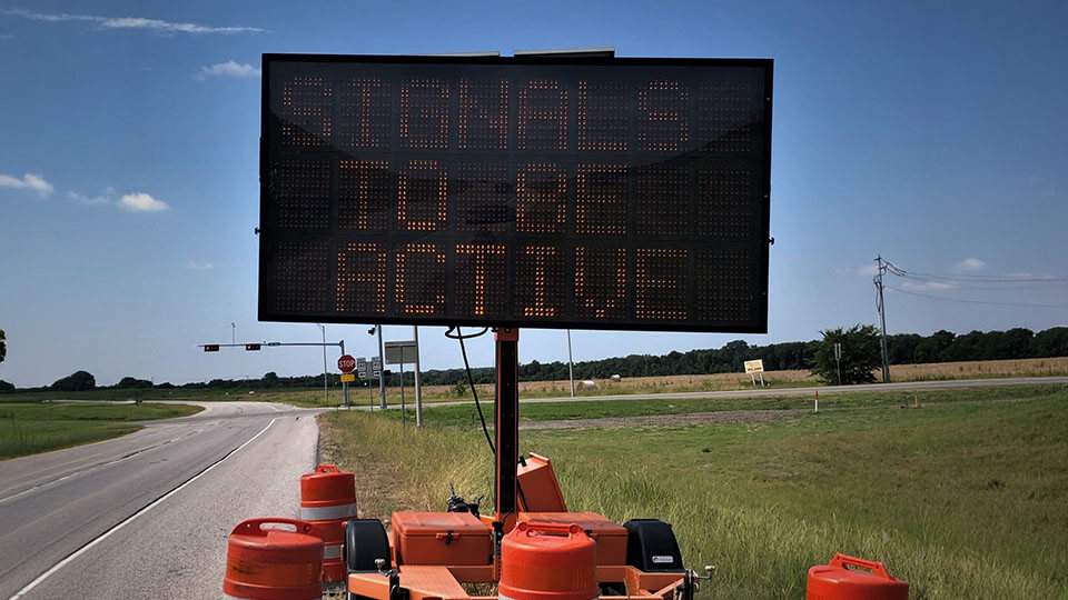 A portable digital roadside message board reading "Signals to Be Active," with orange traffic barrels at its base.