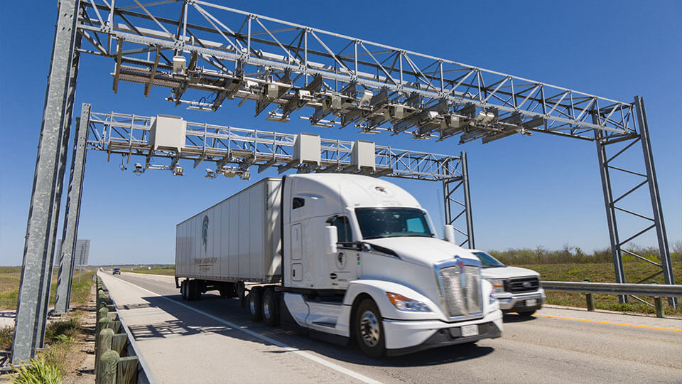 A white semi-truck and pickup truck passing beneath an overhead toll gantry on SH 130.